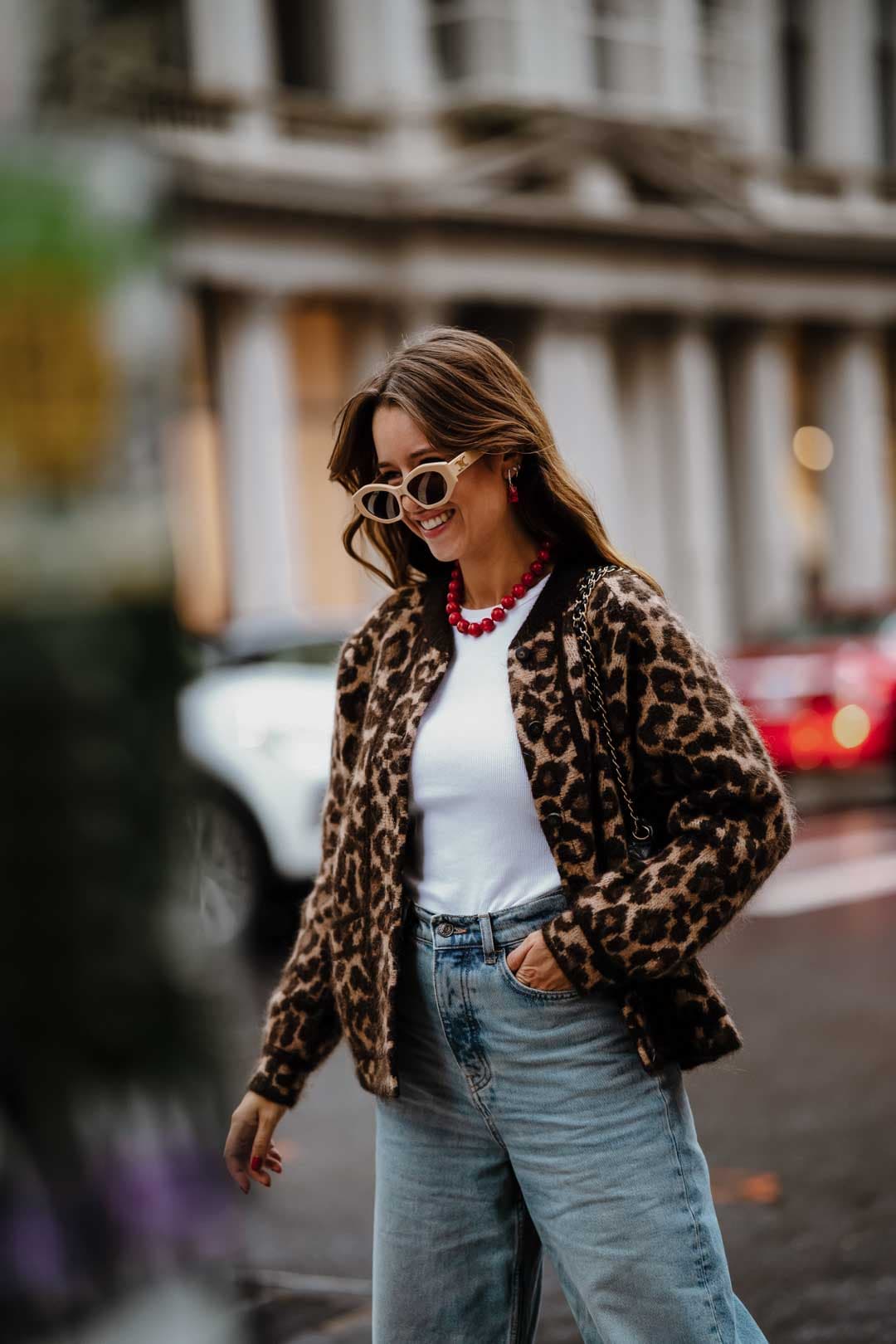 Une femme marche dans la rue avec un gilet léopard, un debardeur blanc, un jeans et des lunettes de soleil