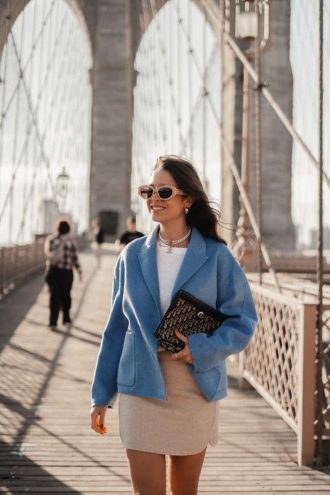 Une femme marche sur un pont à New York. Elle porte une veste bleue, une jupe beige courte, un t-shirt blanc et des lunettes de soleil. Elle a dans les mains une pochettes Dior.