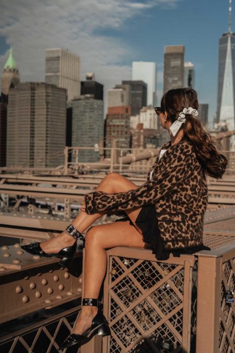 Une femme assise regarde la vue sur New York. Elle est de profil et on peut voir son gilet léopard, sa petite jupe noire et sa paire d'escarpins ouverts noirs avec des détails argentés