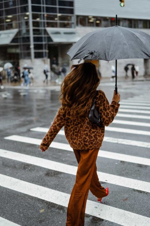 Une femme qui traverse la rue dans une tenue brune. Elle porte un large pull léopard et un pantalon satiné caramel. Ses chaussures sont rouge.