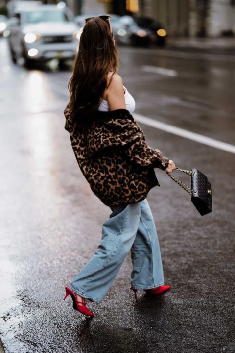 Une femme marche dans la rue de New York et elle porte une tenue d'automne. Il y a un gilet en laine léopard qui tombe de son épaule au dessus d'un débardeur écru. Elle porte un jeans bleu clair et des chaussures à talons rouge. Il y a aussi une paire de lunette beige et un sac chanel noir.