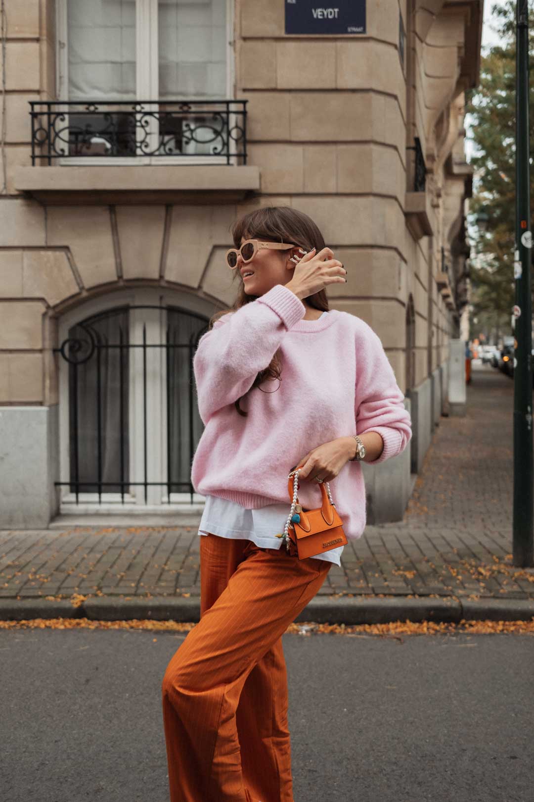 tenue automne American Vintage : pantalon orange rayé, t-shirt blanc oversize, pull en maille rose avec sac Chiquito Jacquemus et baskets Adidas Samba